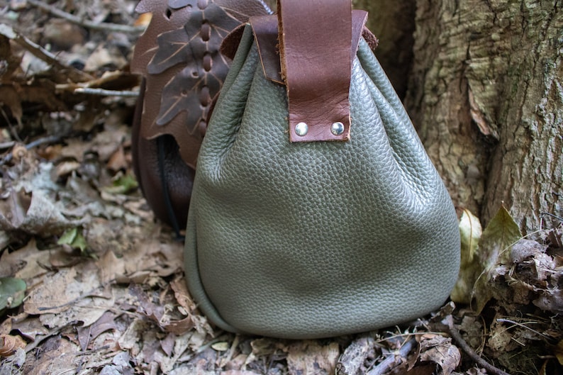 May include: A gray leather pouch with a brown leather strap and metal rivets. The pouch is sitting on a bed of brown leaves.