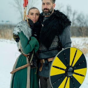 May include: A couple dressed in medieval garb, the woman in a green dress and fur-trimmed cloak, the man in a grey tunic and fur-trimmed cloak, holding a shield and arrows. They are standing in a snowy landscape.