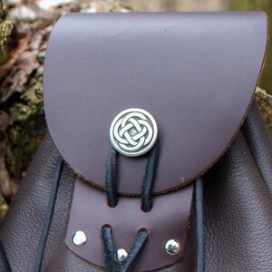 May include: A brown leather bag with a Celtic knot design on the front. The bag has a drawstring closure and a flap with a buckle.