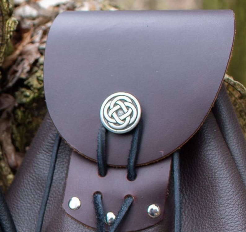 May include: A brown leather bag with a Celtic knot design on the front. The bag has a drawstring closure and a flap with a buckle.