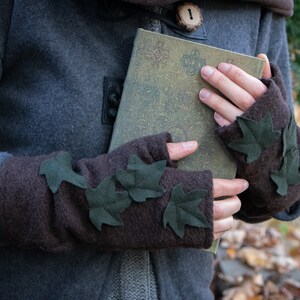 Puede incluir: Guantes sin dedos marrones con hojas de fieltro verde cosidas. Los guantes los lleva una persona que sostiene un libro con una cubierta verde.