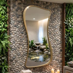 May include: An irregular, organic-shaped mirror with a thin gold frame mounted on a wall covered in small, smooth gray river stones. Below the mirror sits a stack of folded beige towels on a dark wood bench, next to three lit pillar candles.