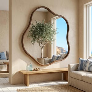 Puede incluir: Un gran espejo de pared con una forma irregular, orgánica y ameboide, con un marco de madera lisa de color marrón oscuro. El espejo refleja un olivo en maceta, una ventana con vistas a un paisaje costero y parte de un sofá con cojines azules.