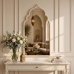 Può includere: Uno specchio da parete con cornice ad arco a sesto acuto che riflette una stanza con divano, una lanterna sospesa e un tavolino. Sotto lo specchio si trova una console bianca con un vaso di vetro con fiori, una piccola lanterna dorata e dei libri.