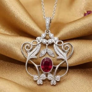 Pode incluir: Um colar com pingente de prata com um design floral. O pingente apresenta uma grande pedra preciosa de rubi oval no centro, rodeada por pedras preciosas claras menores. O colar é exibido em um tecido de cetim dourado.