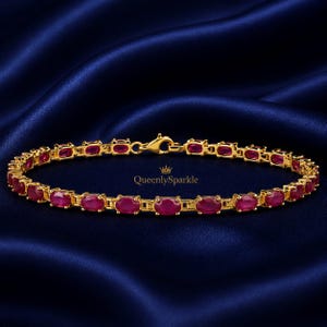Puede incluir: Una pulsera de tono dorado que presenta una serie de piedras preciosas de color rojo intenso con corte ovalado engastadas en eslabones metálicos individuales. Incluye un cierre de mosquetón y el texto QueenlySparkle impreso en letras doradas.