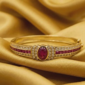 May include: A gold bracelet with a central oval ruby and surrounding diamonds. The bracelet features a band of small rubies and diamonds. The jewelry is displayed on a gold satin surface.