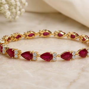 May include: A gold bangle bracelet featuring a repeating pattern of pear-shaped red rubies and small round white diamonds. The stones are set in a continuous circular design, showcasing the vibrant red color of the rubies against the gold metal.