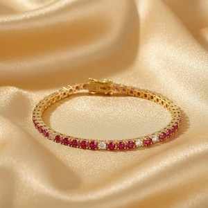 Könnte beinhalten: Ein goldfarbenes Armband mit abwechselnd roten und klaren Edelsteinen. Das Armband liegt auf einer goldenen Satin-Oberfläche. Der Verschluss ist sichtbar und passt zum Goldton des Armbands. Dies ist ein Tennisarmband.