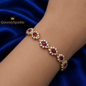 May include: A gold-toned bracelet featuring a series of oval-shaped red stones, each surrounded by a halo of small clear white stones. The text QueenlySparkle is visible in the upper left corner. The design consists of repeating floral-like links.