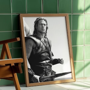 Könnte beinhalten: Gerahmtes Schwarz-Weiß-Foto eines Mannes in einer Lederweste, der ein Schwert hält. Der Holzrahmen lehnt an einer grün gefliesten Wand. Im Vordergrund steht ein Holzstuhl.