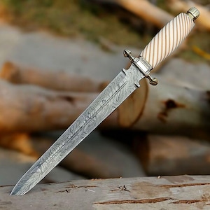 May include: A double-edged dagger featuring a patterned Damascus steel blade with a visible wavy grain. The handle is cream-colored with diagonal grooves and is accented by a polished metallic guard and a matching pommel cap at the end.