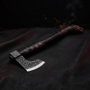 May include: A hand axe featuring a dark brown wooden handle wrapped in black leather cord, ending in a carved wolf head pommel. The metal axe head displays intricate silver-toned Celtic knotwork engravings and runic inscriptions along the blade.