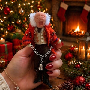 Puede incluir: Una figura navideña en miniatura con una expresión angustiada, que lleva un abrigo rojo con ribete de piel blanca y una cadena plateada con una campana. La figura se sostiene en una mano con esmalte de uñas rojo, con un árbol de Navidad y una chimenea en el fondo.