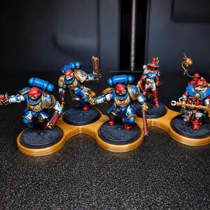 K&ouml;nnte beinhalten: Miniaturfiguren von Space Marines in blauer, roter und goldener R&uuml;stung, auf einzelnen schwarzen Sockeln. Die Figuren sind auf einer goldfarbenen Plattform mit mehreren L&ouml;chern angeordnet. Der dunkle Hintergrund hebt die detaillierte Bemalung hervor.