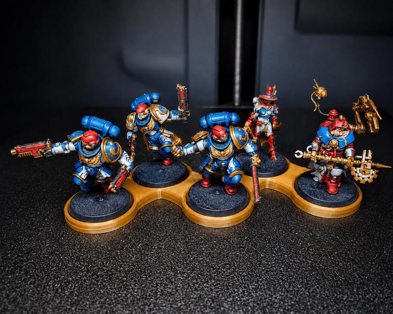 K&ouml;nnte beinhalten: Miniaturfiguren von Space Marines in blauer, roter und goldener R&uuml;stung, auf einzelnen schwarzen Sockeln. Die Figuren sind auf einer goldfarbenen Plattform mit mehreren L&ouml;chern angeordnet. Der dunkle Hintergrund hebt die detaillierte Bemalung hervor.