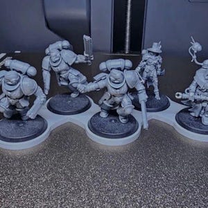 K&ouml;nnte beinhalten: Eine Sammlung grauer Miniaturfiguren, wahrscheinlich f&uuml;r Tabletop-Spiele. Die Figuren sind auf einzelnen schwarzen Sockeln montiert und auf einem hellgrauen Halter angeordnet. Die Figuren stellen gepanzerte Charaktere mit Waffen und Zubeh&ouml;r dar.