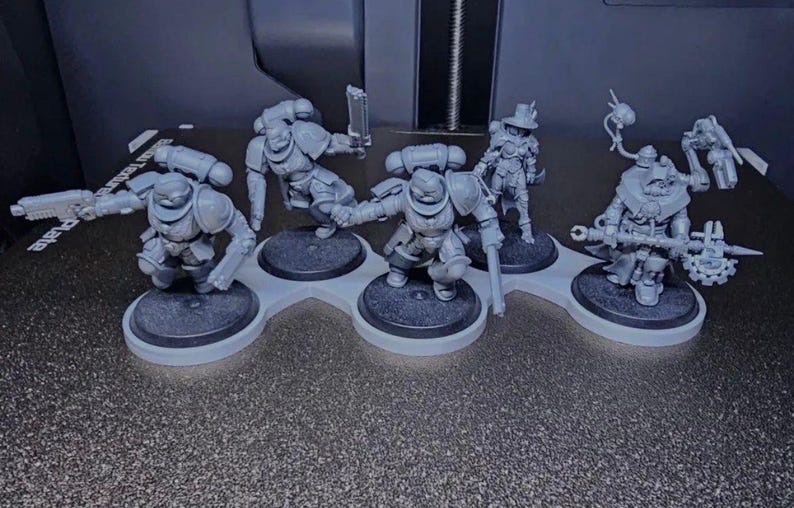 K&ouml;nnte beinhalten: Eine Sammlung grauer Miniaturfiguren, wahrscheinlich f&uuml;r Tabletop-Spiele. Die Figuren sind auf einzelnen schwarzen Sockeln montiert und auf einem hellgrauen Halter angeordnet. Die Figuren stellen gepanzerte Charaktere mit Waffen und Zubeh&ouml;r dar.
