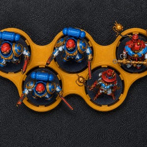 K&ouml;nnte beinhalten: Miniaturfiguren von Space Marines und einem Roboter sind auf einem gelben Tablett angeordnet. Die Space Marines tragen blaue und goldene R&uuml;stungen mit roten Helmen. Der Roboter ist rot und gold. Das Tablett steht auf einer schwarzen Oberfl&auml;che.