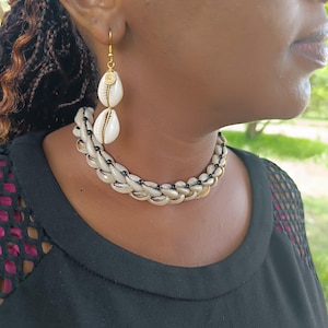 Puede incluir: Primer plano de un collar y pendientes hechos de conchas blancas. El collar tiene un diseño trenzado y los pendientes tienen un detalle de espiral dorada. Las joyas se llevan sobre una camiseta negra.