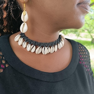 May include: A black top with a black macrame choker necklace and matching earrings. The jewelry features white seashells. The earrings have gold-colored hooks and a spiral design. The background is blurred.
