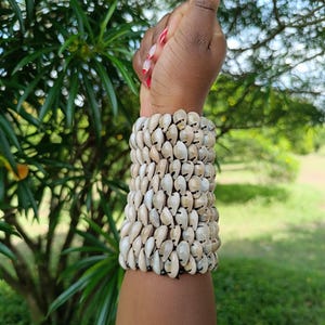 Könnte beinhalten: Ein breites Armband aus zahlreichen kleinen, cremefarbenen Muscheln. Das Armband wird an einem Handgelenk getragen, mit einem Hintergrund aus grünem Laub. Die Nägel der Person sind rot lackiert.