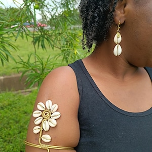 May include: A gold-toned arm cuff and earrings featuring white seashell accents. The arm cuff is designed with a seashell flower and a spiral detail. The earrings have two seashells each. The person is wearing a black tank top.