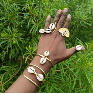 Op de afbeelding: Een hand versierd met goudkleurige sieraden, waaronder ringen en armbanden. De ringen en armbanden hebben witte kaurischelpen. De achtergrond is weelderig groen gebladerte.