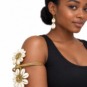 May include: A gold-toned metal arm cuff featuring two flower-shaped designs crafted from white cowrie shells, paired with a matching drop earring consisting of a single gold-toned metal disc and a dangling white cowrie shell on a hook wire.