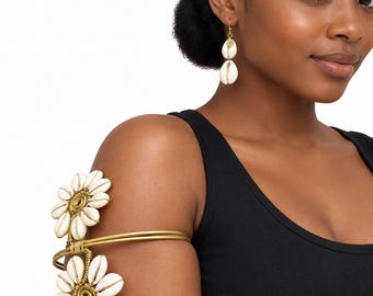 May include: A gold-toned metal arm cuff featuring two flower-shaped designs crafted from white cowrie shells, paired with a matching drop earring consisting of a single gold-toned metal disc and a dangling white cowrie shell on a hook wire.