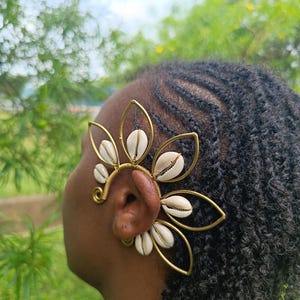 Könnte beinhalten: Ein goldfarbener Ohrschmuck mit floralem Design und weißen Muscheln. Der Ohrschmuck wird am Ohr getragen. Der Hintergrund ist grünes Laub.