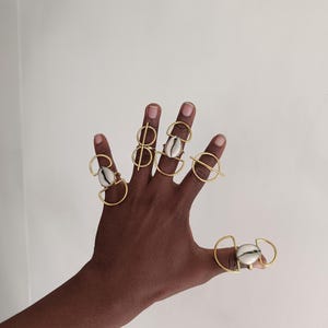 Puede incluir: Una mano que muestra varios anillos dorados. Cada anillo presenta un diseño único, algunos con formas geométricas y otros con un acento de concha. Los anillos están colocados sobre un fondo blanco liso.