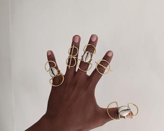 May include: A hand displaying several gold-toned rings. Each ring features a unique design, some with geometric shapes and others with a seashell accent. The rings are set against a plain white background.