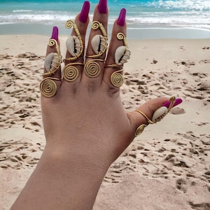 May include: A hand adorned with long, magenta fingernails and gold-coloured wire rings. Each ring features a spiral design and a small, white seashell. The background shows a sandy beach and the sea.