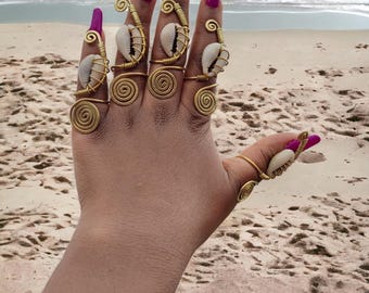 May include: A hand with long, magenta-colored fingernails and gold-colored wire rings. Each ring features a spiral design and a small, white seashell. The background shows a sandy beach and the ocean.
