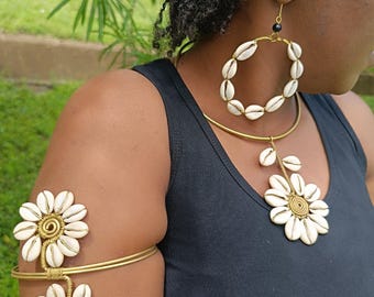 May include: A woman wearing a matching jewelry set. The set includes a necklace, earrings, and an arm cuff. The jewelry features white seashells and gold-colored wire. The necklace has a flower pendant, and the earrings are hoop-shaped.