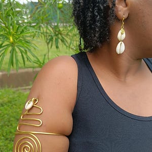 May include: A gold-toned arm cuff with a spiral design and a seashell accent, paired with matching seashell earrings. The jewelry is worn on a person's arm and ear, with a black tank top in the background.