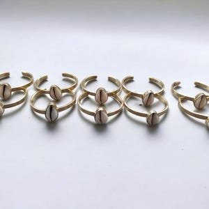 Puede incluir: Pulseras de puño de metal dorado, cada una adornada con una sola concha en forma de lágrima, de color blanquecino y beige. Las pulseras están dispuestas en una fila ordenada, mostrando su diseño simple pero elegante.