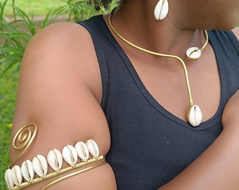 May include: Gold-toned jewelry set featuring cowrie shells. The set includes a necklace, earrings, and an arm cuff. The arm cuff has a spiral design. The jewelry is worn against a black top, with a green background.