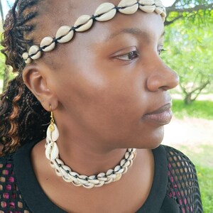 May include: A cream seashell jewellery set including a headband, necklace, and earrings. The headband and necklace are made of cream-coloured seashells strung together with black string. The earrings have a gold-coloured hook and a seashell design.