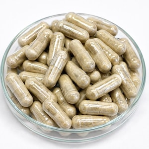May include: A clear glass dish filled with numerous beige capsules. The capsules are cylindrical with rounded ends, and appear to contain a light-colored powder. The dish is set against a white background, highlighting the capsules' texture and form.