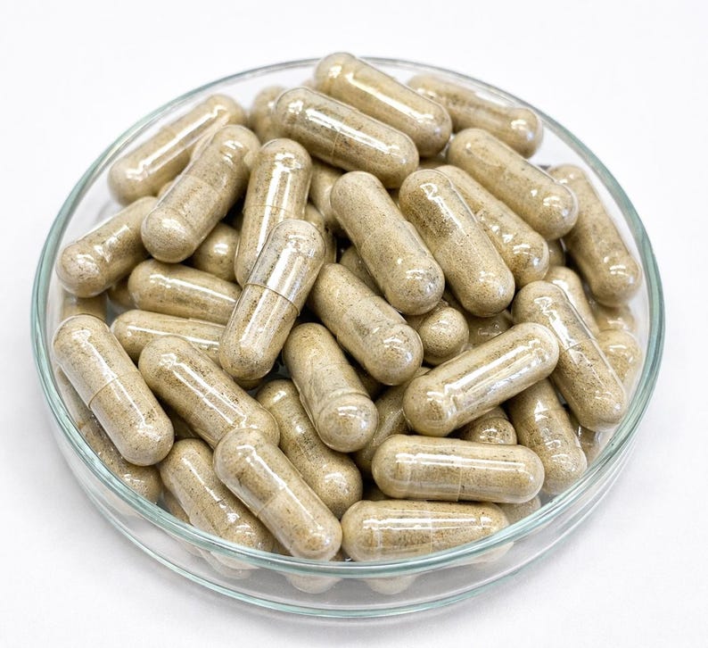 May include: A clear glass dish filled with numerous beige capsules. The capsules are cylindrical with rounded ends, and appear to contain a light-colored powder. The dish is set against a white background, highlighting the capsules' texture and form.