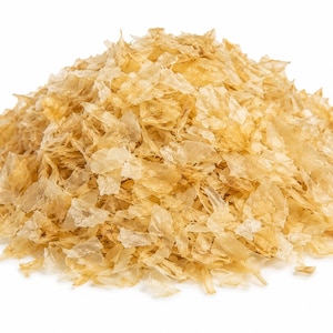 Puede incluir: Un montón de escamas translúcidas de color marrón dorado claro con bordes irregulares y dentados. El material parece fino, seco y quebradizo, con una textura superpuesta que crea un aspecto variado y semitransparente en todo el montón.