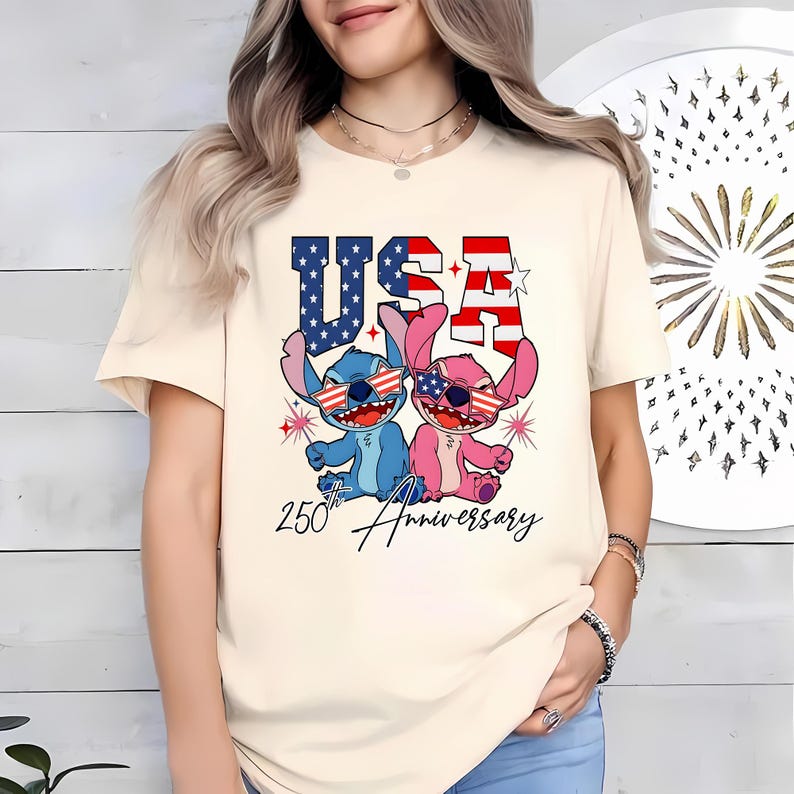 May include: A cream-coloured t-shirt with a graphic of two cartoon characters wearing sunglasses with a USA flag design. The word "USA" is in red and blue, with "250th Anniversary" below.