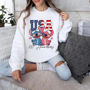 May include: White sweatshirt with a graphic of two cartoon characters wearing sunglasses with the American flag design. The word "USA" is above the characters, and "250th Anniversary" is written below.