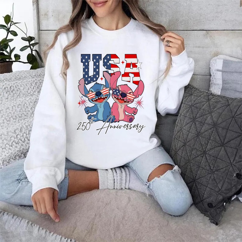 May include: White sweatshirt with a graphic of two cartoon characters wearing sunglasses with the American flag design. The word "USA" is above the characters, and "250th Anniversary" is written below.
