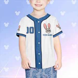 May include: White and blue baseball jersey featuring a cartoon rabbit design. The jersey has a blue collar, sleeves, and button placket. The number "10" is on the front, and the text "YOUR NAME" is below the rabbit graphic. Worn by a child.