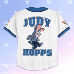 May include: White baseball jersey with blue trim and the name "JUDY HOPPS" in blue block letters. A cartoon image of Judy Hopps, a gray rabbit, is featured on the back. The jersey has short sleeves and a button-up front.