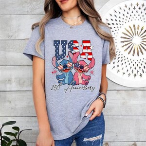 May include: A light grey t-shirt with a graphic of two cartoon characters wearing sunglasses in the style of the American flag. The word "USA" is above the characters, and "250th Anniversary" is written below.