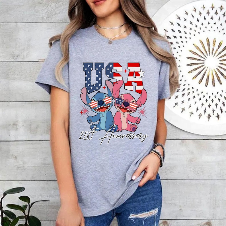 May include: A light grey t-shirt with a graphic of two cartoon characters wearing sunglasses in the style of the American flag. The word "USA" is above the characters, and "250th Anniversary" is written below.